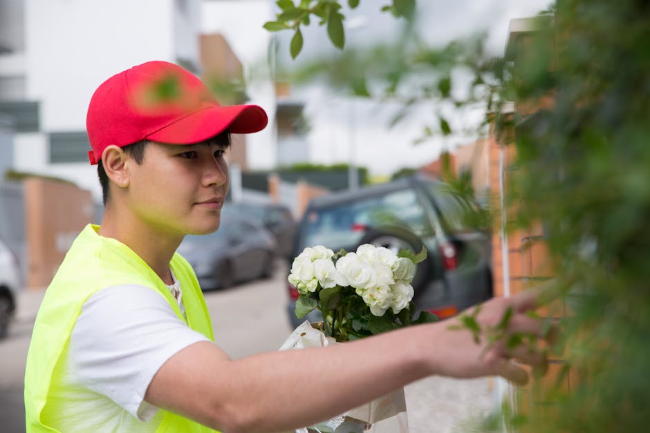 A delivery person wearing a red cap and a neon yellow safety vest is shown holding a bouquet of white hydrangeas while standing outdoors on a residential street. The bouquet is wrapped in white paper