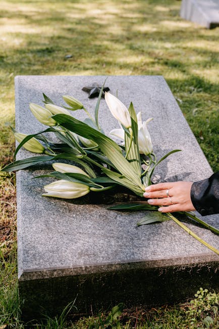 A person dressed in a black leather jacket places a bouquet of white lilies with long, green leaves and sturdy stems onto a polished grey granite gravestone. The bouquet, freshly cut, features several