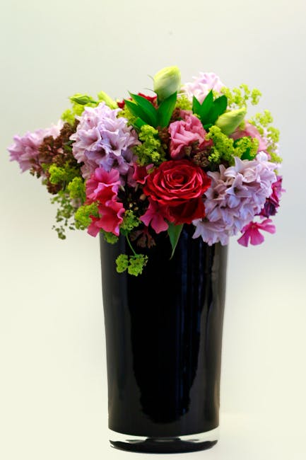A tall, elegant black glass vase holding a vibrant floral arrangement featuring pink and lavender hydrangeas, red roses, white tulips, and green foliage. The bouquet is lush and full, with the flowers