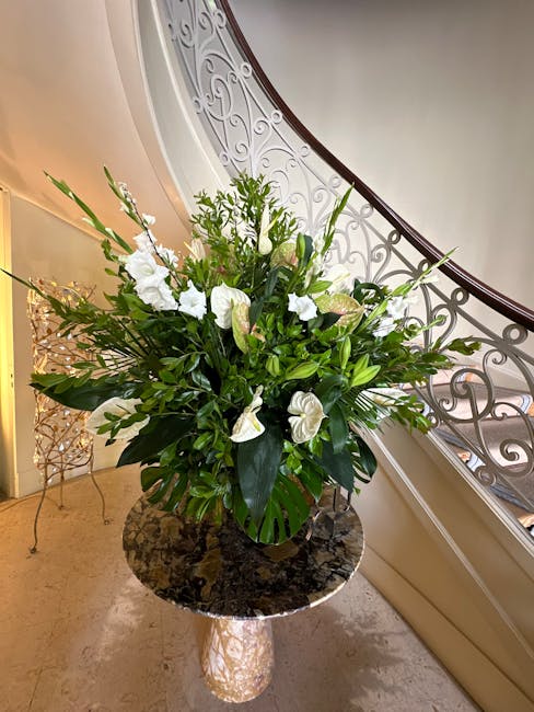A large, elegant floral arrangement in a clear glass vase, featuring white lilies, anthuriums, and various lush green foliage including broad leaves and delicate fronds. The bouquet is placed on a pol