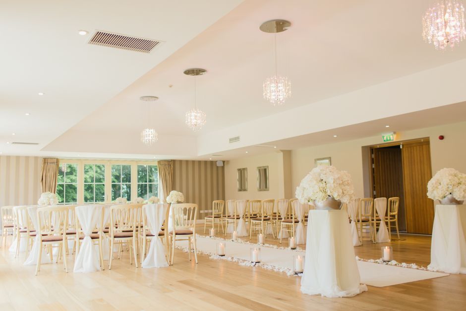 A spacious, well-lit indoor venue decorated for a wedding ceremony with elegant floral arrangements. The room features large windows allowing natural light to illuminate the space, with cream-colored