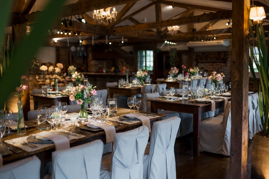 An elegantly arranged wedding reception dining area featuring several rectangular wooden tables decorated with floral centrepieces composed of pink and white roses, green foliage, and small filler flo