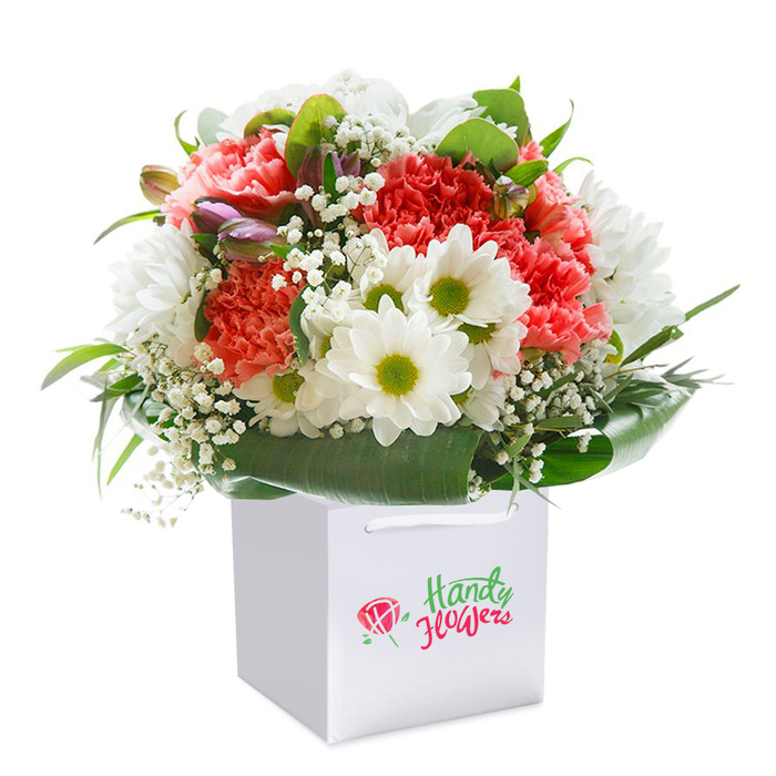 A lush, exuberant floral arrangement stands tall and full within a smart white square gift bag, its beauty unmistakably crafted by the expert hands of Noak Hill's local florists. The composition is anchored by clusters of soft coral and peach-hued carnations, distinguished by their frilled, almost feathery petals that invite touch and admiration. Bright red carnations add boldness, weaving through the bouquet like ribbons of excitement. In gentle contrast, snow-white daisies open their petals toward the light, each one crowned by a lemon-lime center that draws the eye and brings a fresh, optimistic mood. Delicate tendrils of baby's breath drift among the larger blooms, their tiny blossoms forming a misty veil that lends softness and dimension. Peeking discreetly from the left side are a handful of closed buds, tinged in purple, hints of future blossoms and moments yet to bloom-a subtle nod to the ever-renewing cycle of floral artistry. Embracing the flowers is an abundance of deep, shiny green foliage, unfurling in smooth, broad arcs to form a rich natural collar around the arrangement. The bouquet is perfectly poised in its white gift bag, complete with robust ribbon handles for effortless carrying to homes across Noak Hill, whether just off Noak Hill Road or near the edges of the surrounding countryside. Set against a seamless white background, every aspect of the arrangement glows with clarity and pride. This presentation radiates thoughtfulness, quality, and the unique elegance that marks every special moment celebrated in Noak Hill.