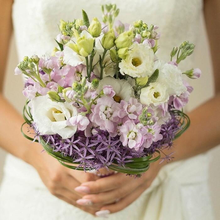 A beautifully arranged bridal bouquet is gently held by a bride in the heart of Noak Hill, her hands softly cupping the stems just below a cascade of creamy whites, pastel pinks, and rich purples. The dominant white Lisianthus blossoms, each with intricate ruffled petals and delicate yellow-green eyes, evoke purity and quiet sophistication-an elegant match for the lace-trimmed gown, just visible in the blurred background. Nestled amongst the Lisianthus are plump tufts of pale lilac and rosy pink stock, their myriad petals providing both texture and subtle fragrance that lingers in the spring air. Sprigs of soft green buds lend a breath of freshness and anticipation, while spiky, deep purple Allium at the bouquet's base introduces dramatic contrast and architectural intrigue-a flourish inspired perhaps by the vibrant community gardens blooming across Noak Hill in early summer. Above this, fine threads of dark bear grass swirl in delicate loops, thoughtfully placed for a touch of modern drama and structural grace. The overall aesthetic is at once romantic and refined, a floral tribute to new beginnings created for the most special of occasions-a perfect keepsake image for a wedding, engagement, or styled shoot in Noak Hill, crafted with evident passion by skilled local hands.